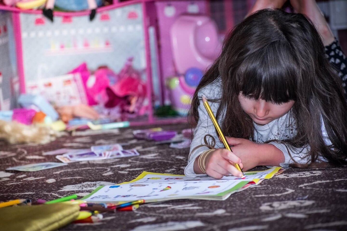 A young girl with dark brown hair is lying on the floor and writing on a worksheet. In the background are lots of children's toys