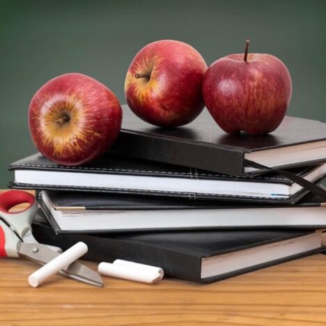 A stack of black notebooks on a desk, with 3 red apples on top. Some sticks of white chalk and a pair of scissors are next to it