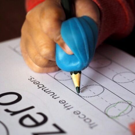 A child's hand holding a pencil using an ergonomic pencil grip. They are writing on a piece of paper the letter O