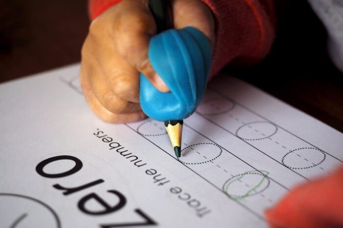 A child's hand holding a pencil using an ergonomic pencil grip. They are writing on a piece of paper the letter O