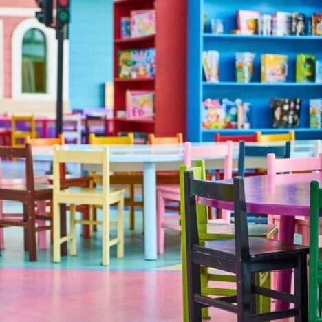 A colourful children's nursery or library. Brightly coloured seats of different colours are around a table. Bookshelves are also brightly coloured.