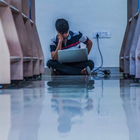 A man sitting on the floor of a library working on a laptop that is plugged in