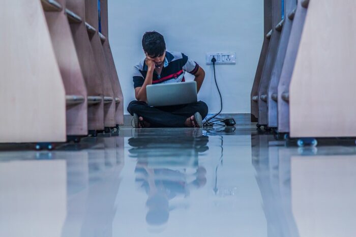 A man sitting on the floor of a library working on a laptop that is plugged in