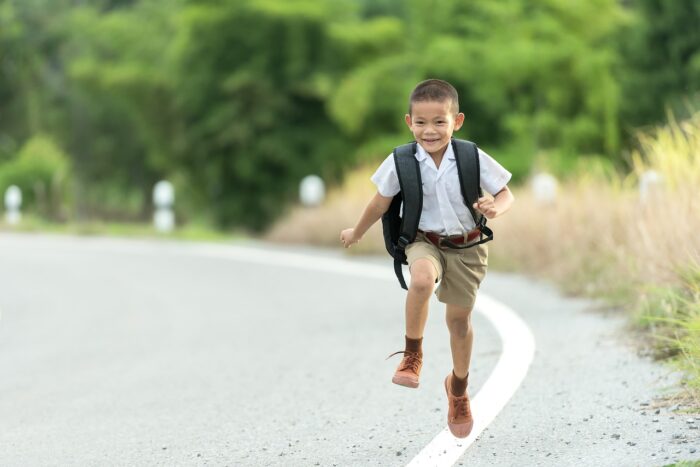 A boy running along a road. He is in school uniform and has a backpack on. There is a big smile on his face.