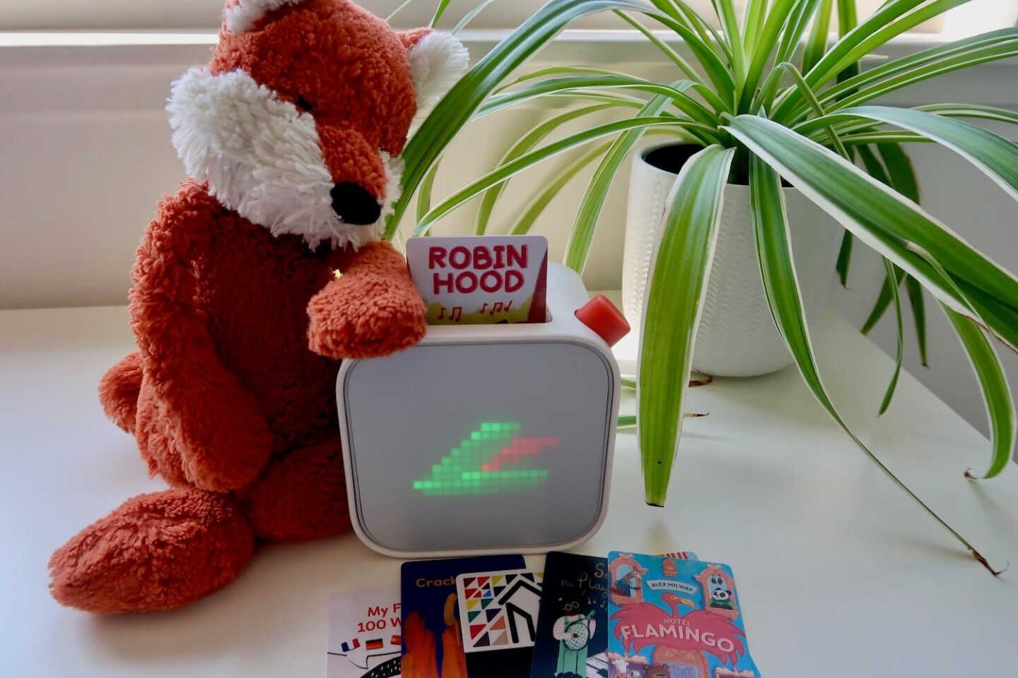A Yoto player with a green hat with a red feather in it, of the style of Robin Hood. A Robin Hood card is in the player, and some more Yoto cards lay in front. A fox soft toy sits next to it, with a spider plant in the background