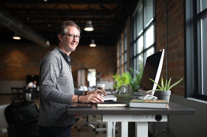 A man sitting at a desk in an office with high ceilings and big windows. He is smiling at the camera with his hand on his mouse